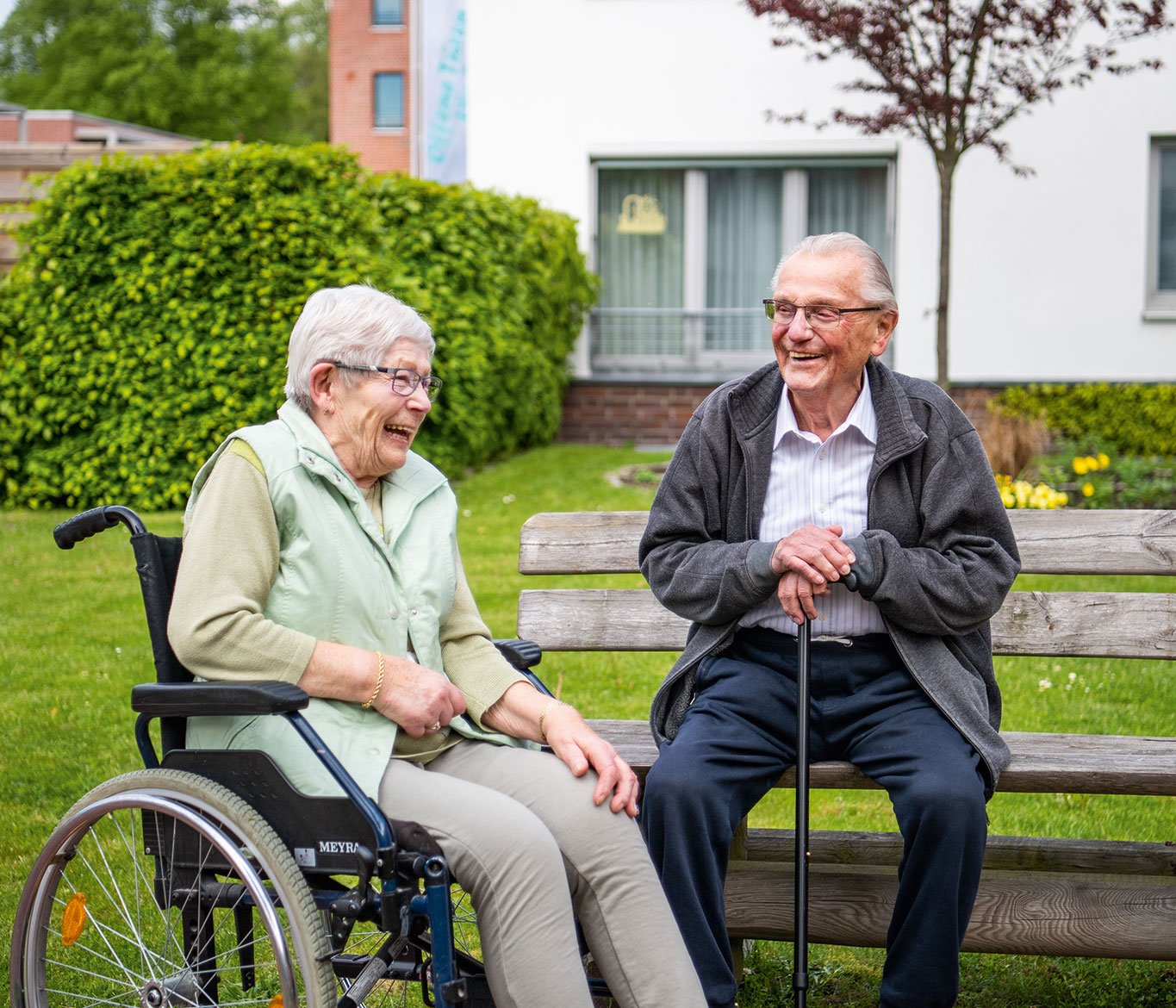 Eine Seniorin im Rollstuhl lacht mit einem Senior vor einer Einrichtung der Seniorenpflege der Vorwerker Diakonie.