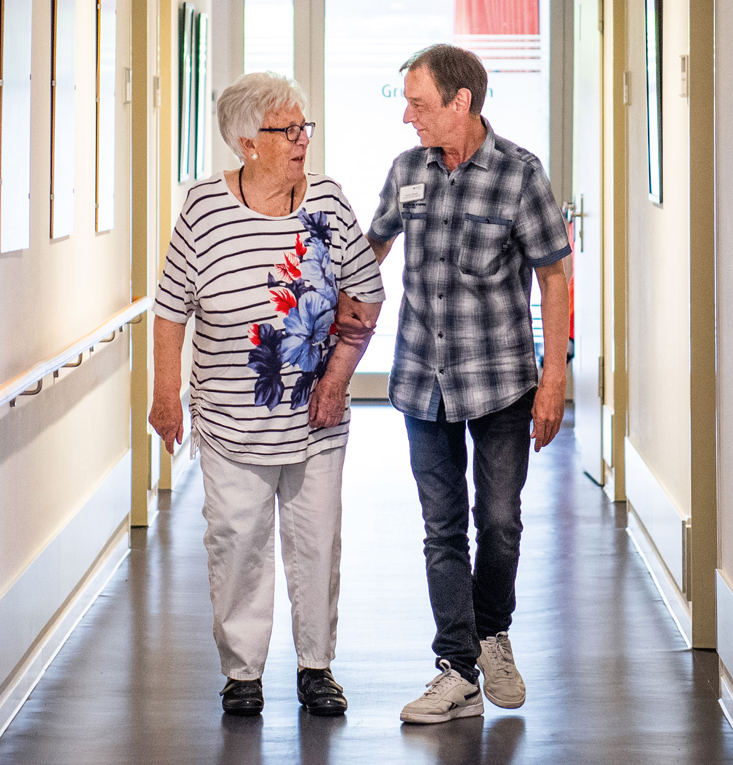 Ein Pfleger geht mit einer Seniorin in einer Einrichtung der Seniorenpflege den Gang entlang.