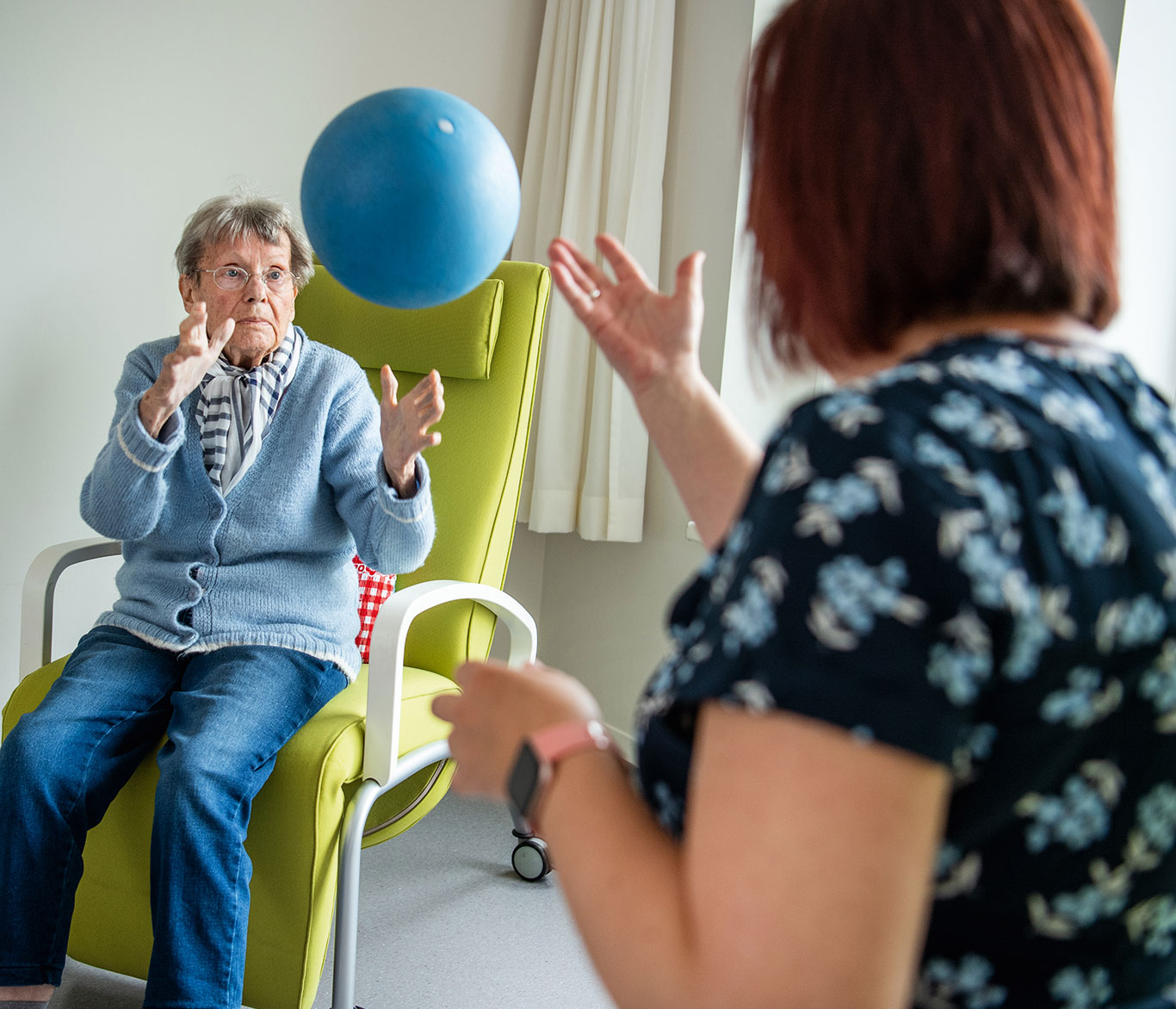 Eine Pflegerin wirft einer im Sessel sitzenden Seniorin in der Tagespflege der Vorwerker Diakonie einen blauen Ball zu.
