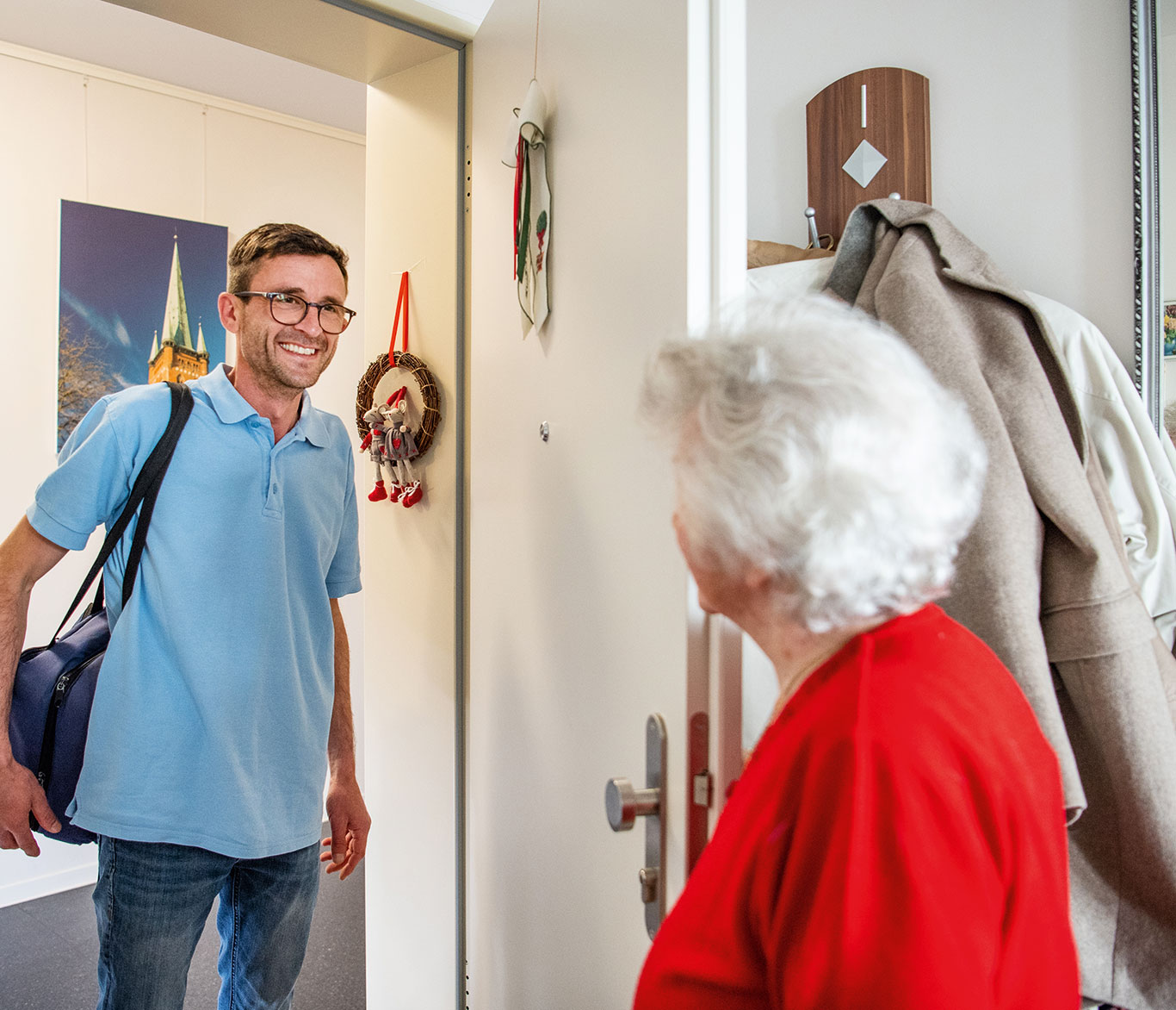 Ein Pfleger für die ambulante Pflege begrüßt eine Seniorin an ihrer Wohnungstür in ihrem Zuhause.