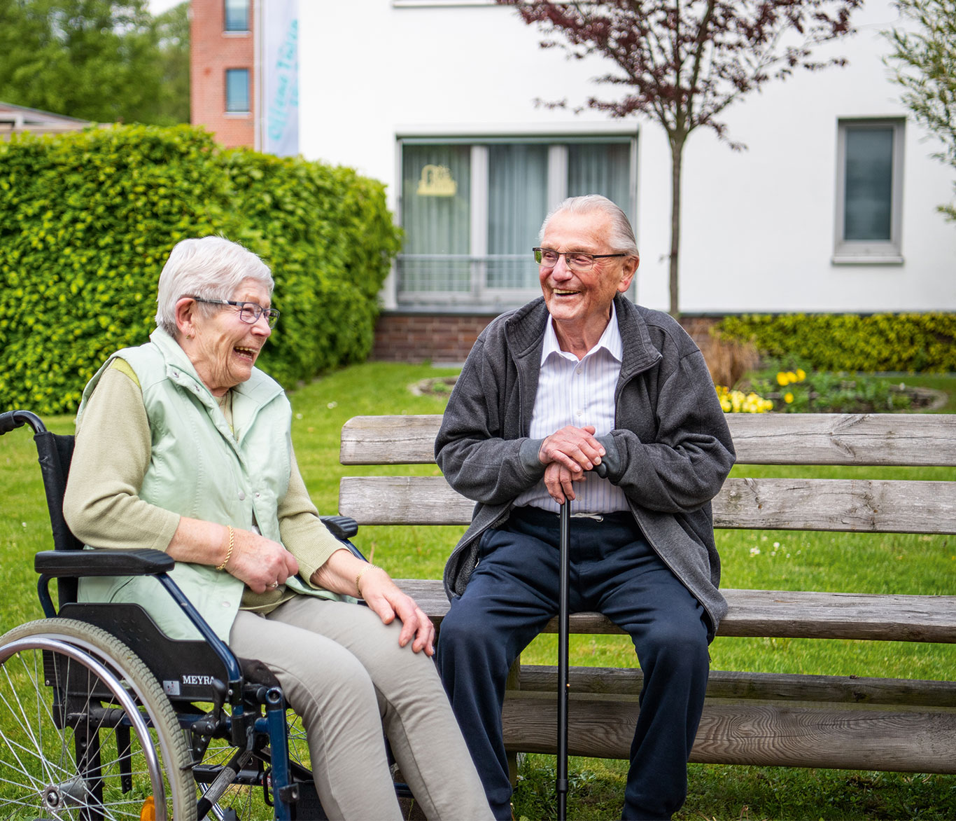 Eine Seniorin im Rollstuhl lacht mit einem Senior vor einer Einrichtung der Seniorenpflege der Vorwerker Diakonie.