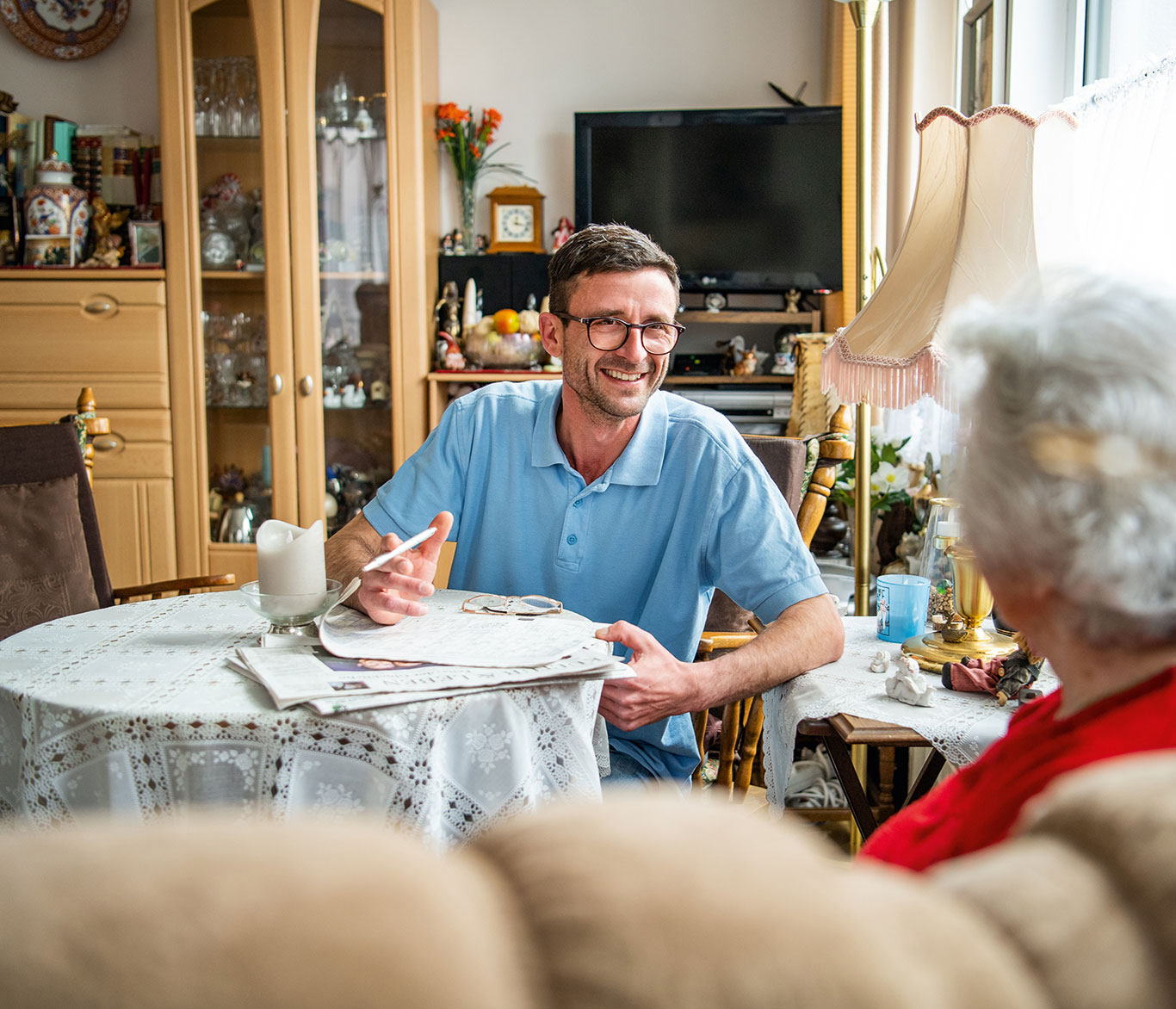 Pfleger besucht bei der ambulanten Pflege eine Seniorin und unterhält sich mit ihr in einer Sitzecke in ihrem Zuhause.